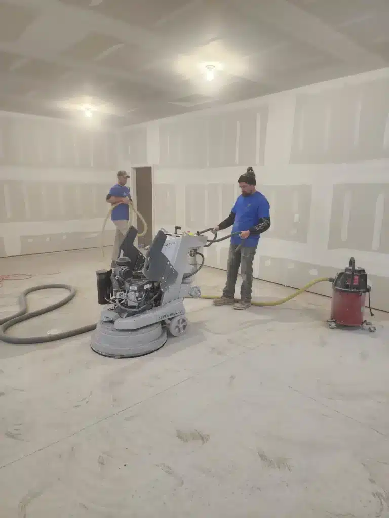 Two persons are working in a drywalled room with a concrete floor. One operates a large floor grinder, while the other handles a vacuum system.