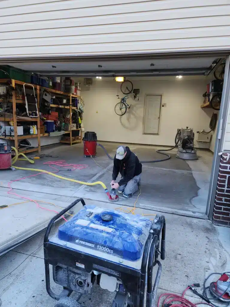 A person is kneeling on a garage floor, working with a tool near a generator. Shelves with various items and bicycles are also visible.