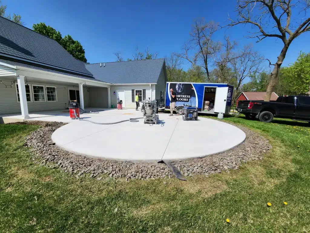 A fresh concrete driveway is being constructed outside a house with a person working, concrete equipment, a trailer labeled "FORTRESS FLOORS", and two trucks parked nearby.