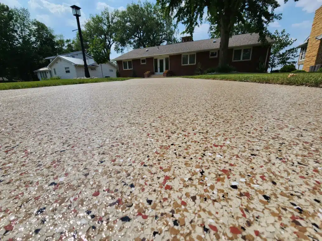 A low-angle view of a textured driveway leading to suburban houses, with clear skies and green trees in the background. No people are visible.