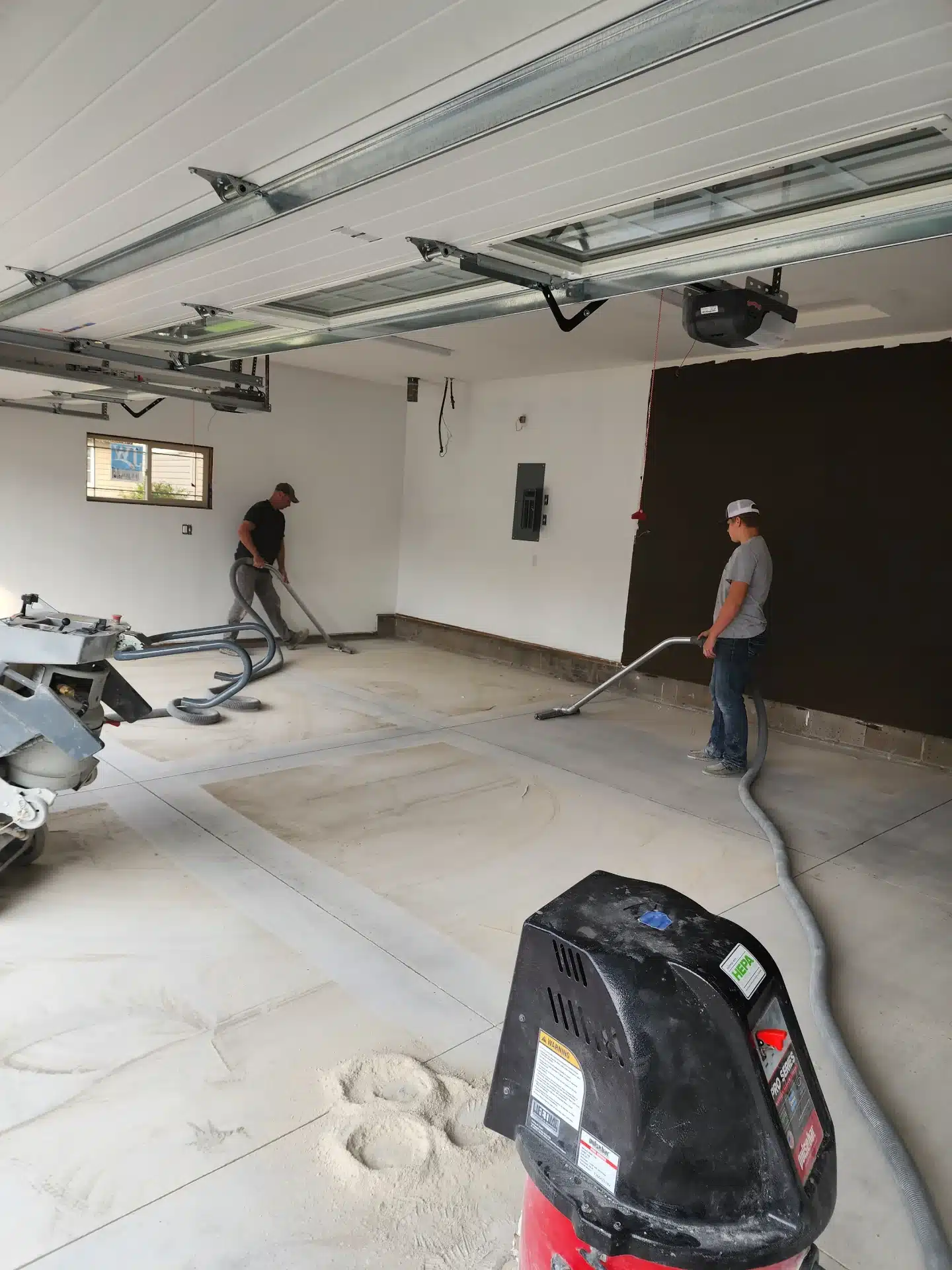 Two people are working in a garage, using machinery to polish a concrete floor. There's a vacuum cleaner and a power cord on the foreground.