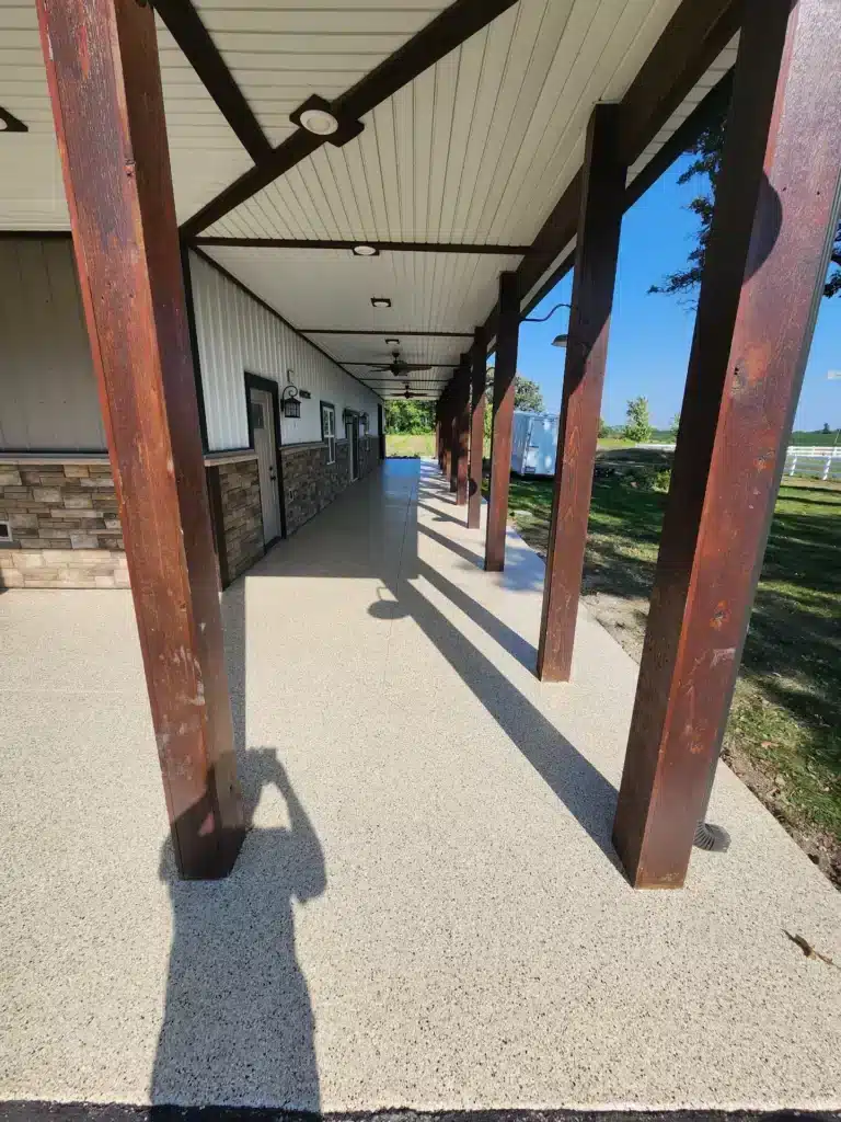 A covered walkway with brown supports and a white ceiling. Sunlight casts the shadow of a person taking the photo, clear blue sky visible.