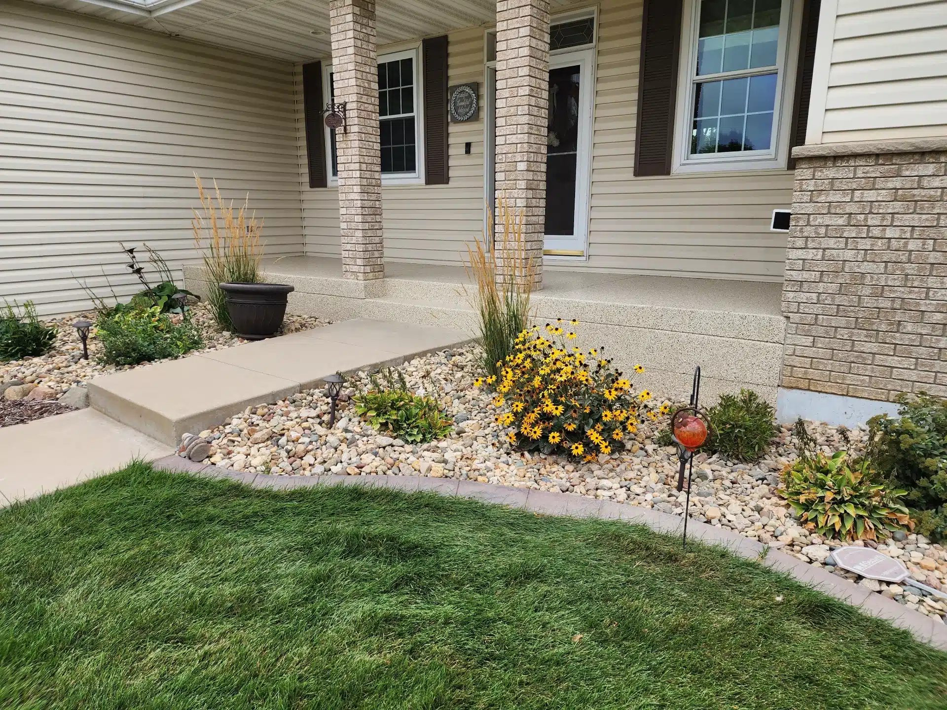 A well-kept garden in front of a house with green grass, a pebble bed, potted plants, yellow flowers, and a decorative glass sphere.