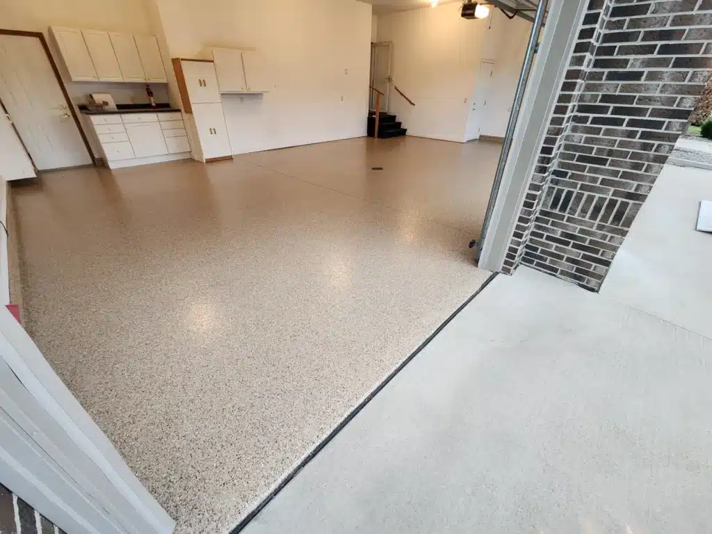 This is an empty garage interior with a speckled epoxy floor, white storage cabinets, a brick wall on the right, and a staircase in the back.