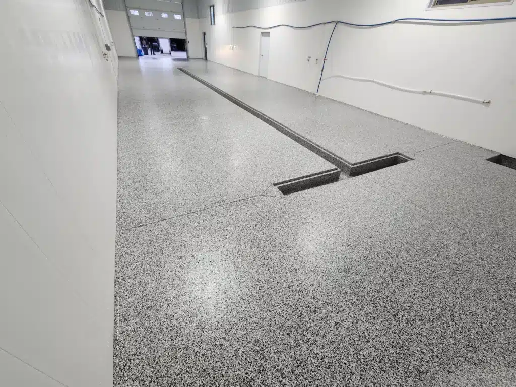 An empty industrial indoor space with a shiny speckled epoxy floor, white walls, some cables, and a distant open doorway leading to a brighter area.