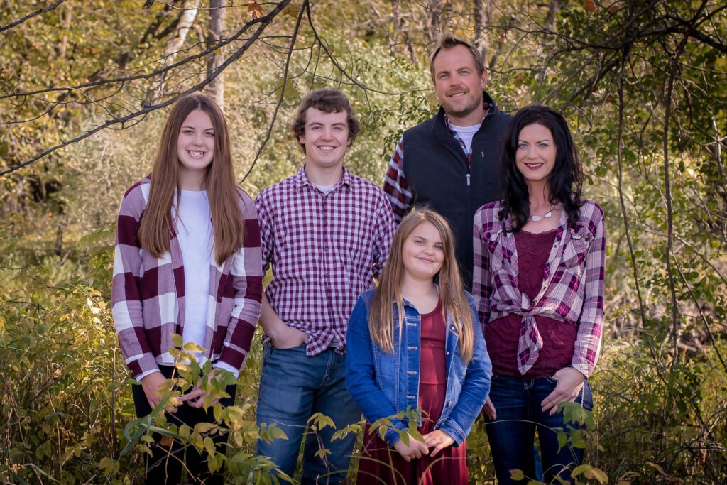 A family of five poses outdoors amidst lush greenery, all wearing plaid shirts, smiling warmly at the camera on a sunny day.